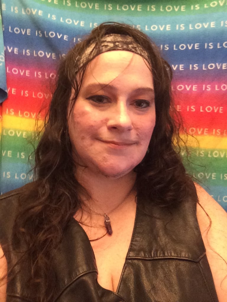 Image of J. Yve Brown. A person wearing a leather vest and headband in front of a rainbow "Love is Love backdrop.